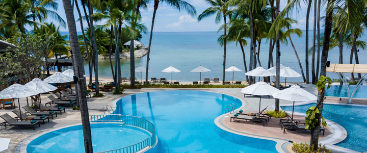 Schräg von oben fotografierter, leuchtend blauer Pool mit Liegen und Sonnenschirmen; dahinter das Meer, Palmen ragen aus der Poolanlage hervor.